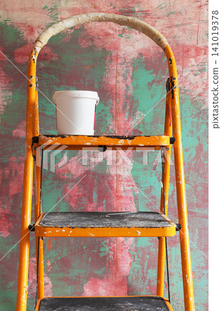 Yellow paint-splattered step ladder with white paint bucket standing against colorful textured wall during home renovation. Interior painting, DIY remodeling, and decoration concept 141019378