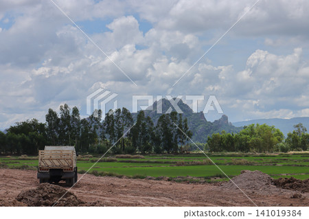 Rural landscape with truck dirt road green field mountain cloudy sky tree line farm open field countryside construction site Rural landscape with truck dirt road green field mountain cloudy sky tree line farm open field countryside construction site 141019384