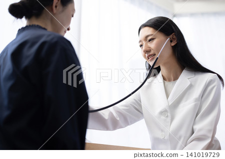 A young Japanese female doctor using a stethoscope during an examination. A young Japanese female doctor using a stethoscope during an examination. 141019729