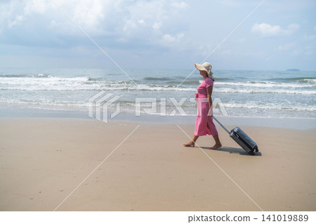 Young woman in pink dress and straw hat carrying a grey suitcase walking along a wide flat beach with sea on one side and open sky above. Coastal travel concept 141019889