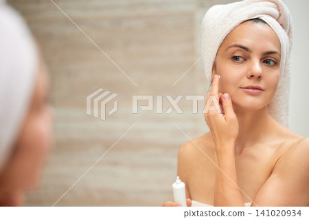 Woman in a towel applying facial cream to her cheek in the bathroom, focusing on her morning beauty and wellness routine Woman in a towel applying facial cream to her cheek in the bathroom, focusing on her morning beauty and wellness routine 141020934