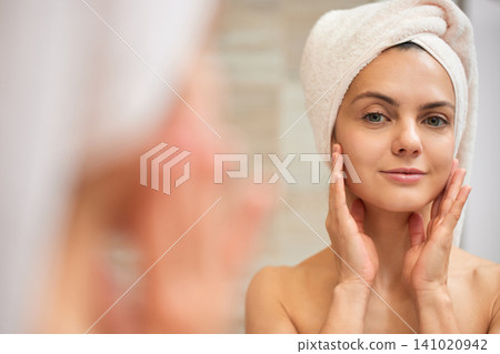 Woman in a towel reflecting in a mirror, applying skincare product to her cheeks for beauty and wellness routine 141020942