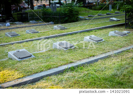 Rows of flat grave markers on grassy cemetery grounds with soft sunlight, trimmed hedges, stone pathways, and calm memorial setting across maintained lawn, with grave, marker, cemetery, memorial Rows of flat grave markers on grassy cemetery grounds with soft sunlight, trimmed hedges, stone pathways, and calm memorial setting across maintained lawn, with grave, marker, cemetery, memorial 141021091