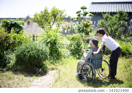 Home care: An elderly person in a wheelchair and a woman in the garden of a home (back view). Home care: An elderly person in a wheelchair and a woman in the garden of a home (back view). 141021400