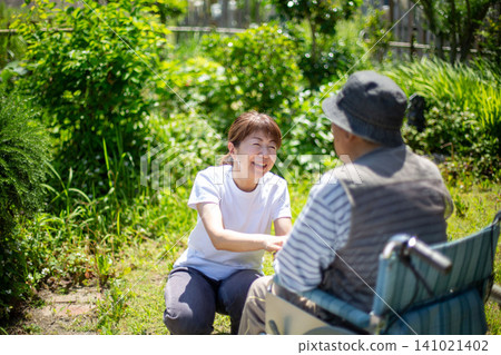Home care: An elderly person in a wheelchair and a woman in the garden of a home (back view). Home care: An elderly person in a wheelchair and a woman in the garden of a home (back view). 141021402