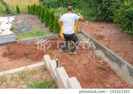 A construction worker in a white t-shirt pushing a yellow wheelbarrow with soil during backyard renovation and landscaping works near a new concrete retaining wall. High-angle view of a builder A construction worker in a white t-shirt pushing a yellow wheelbarrow with soil during backyard renovation and landscaping works near a new concrete retaining wall. High-angle view of a builder 141021481