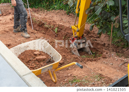 excavator actively digging soil at a construction site, surrounded by a wheelbarrow and greenery, captured in a side view with vibrant yellow and red tones, creating a productive and clean. 141021505