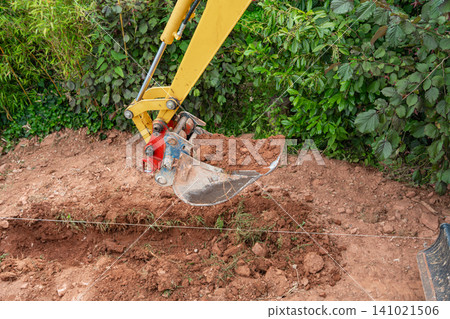 Close-up of a yellow mini excavator steel bucket lifted up and filled with brown soil directly above a dug trench. Perfect for earthmoving and site development concepts. Mini digger bucket full of Close-up of a yellow mini excavator steel bucket lifted up and filled with brown soil directly above a dug trench. Perfect for earthmoving and site development concepts. Mini digger bucket full of 141021506