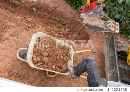Top-down close-up shot of a mini excavator bucket loading fresh brown soil into a metal wheelbarrow on a construction site. High-quality image for earthwork services. Mini digger bucket filling a Top-down close-up shot of a mini excavator bucket loading fresh brown soil into a metal wheelbarrow on a construction site. High-quality image for earthwork services. Mini digger bucket filling a 141021508