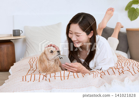 A young woman with a smile lies on her bed, looking at her smartphone while stroking her dog. 141021828