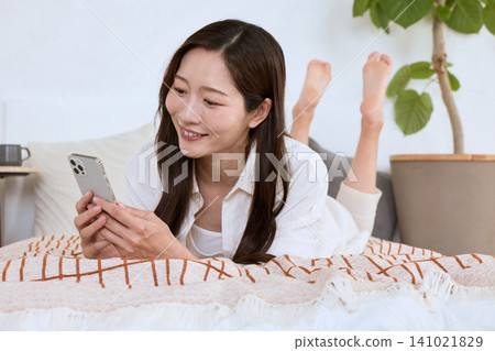 A young woman with a smile is lying on a bed looking at her smartphone. 141021829