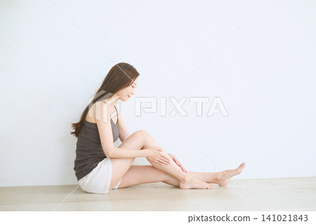 A young woman sitting in front of a white wall, taking care of her legs. A young woman sitting in front of a white wall, taking care of her legs. 141021843