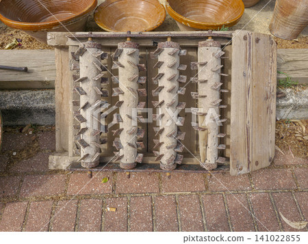 Wooden tilling tool with iron rollers sits on the ground near clay pots. This scene shows traditional farming practices in a rural area. The tool is ready for use during planting season. Wooden tilling tool with iron rollers sits on the ground near clay pots. This scene shows traditional farming practices in a rural area. The tool is ready for use during planting season. 141022855