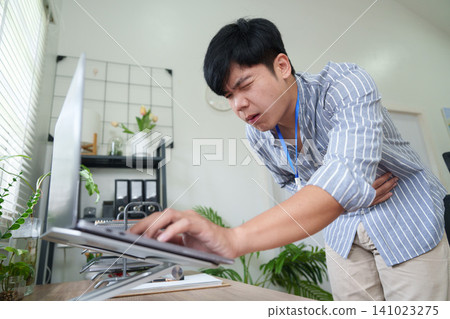 Asian man clutching stomach in pain while working on laptop at desk Asian man clutching stomach in pain while working on laptop at desk 141023275