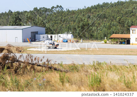Small aircraft parked on a helipad near a hangar and fuel station, surrounded by trees and grass at a rural airfield in a sunny environment Small aircraft parked on a helipad near a hangar and fuel station, surrounded by trees and grass at a rural airfield in a sunny environment 141024337