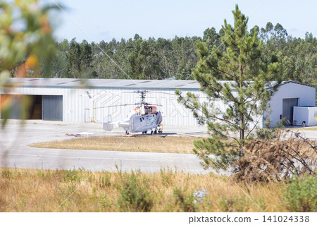 Helicopter parked on a tarmac near a large hangar surrounded by trees and grass, showcasing a clear blue sky in the background Helicopter parked on a tarmac near a large hangar surrounded by trees and grass, showcasing a clear blue sky in the background 141024338
