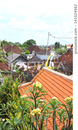 Tropical Bali village rooftops with frangipani blossoms and temple shrines beneath cloudy sky, framed by lush greenery, terracotta tiles, traditional ornament, and distant horizon 141024982