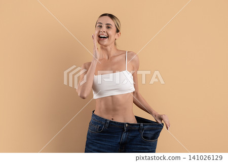 Young beautiful woman laughing and showing oversized jeans on beige studio background Young beautiful woman laughing and showing oversized jeans on beige studio background 141026129