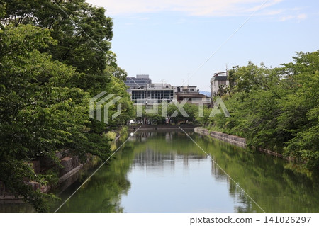 A townscape with green trees, buildings, and a canal reflecting the sky on its surface. A townscape with green trees, buildings, and a canal reflecting the sky on its surface. 141026297