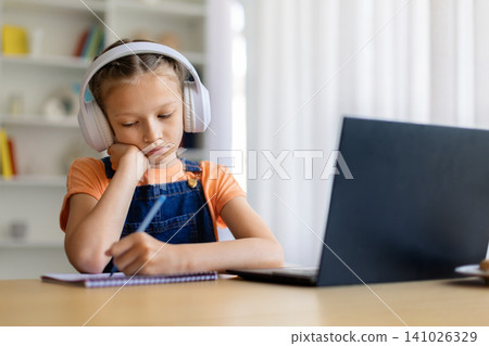 Teen girl in white headphones writes notes beside laptop at home Teen girl in white headphones writes notes beside laptop at home 141026329