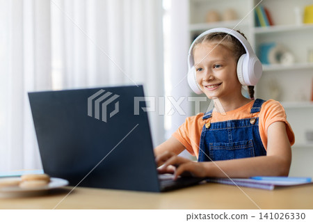 Teen girl wearing white headphones smiling while typing on laptop at home Teen girl wearing white headphones smiling while typing on laptop at home 141026330