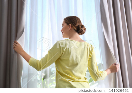 Woman opening window curtains at home in morning Woman opening window curtains at home in morning 141026461