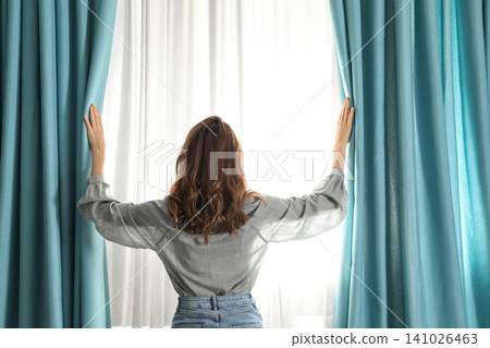 Woman opening window curtains at home in morning, back view Woman opening window curtains at home in morning, back view 141026463