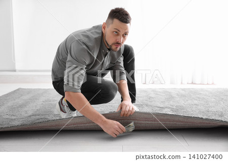 Worried man hiding money under carpet indoors. Financial savings Worried man hiding money under carpet indoors. Financial savings 141027400