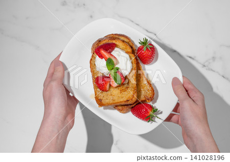 Hands holding a plate of brioche toasts with strawberry and whipped cream, drizzled with honey Hands holding a plate of brioche toasts with strawberry and whipped cream, drizzled with honey 141028196