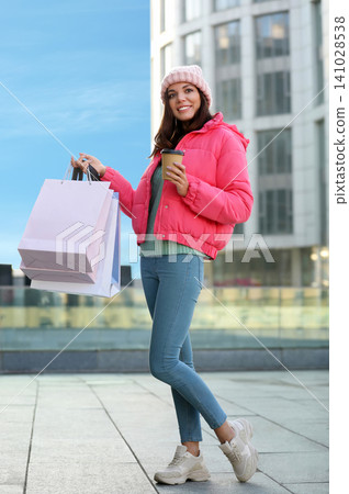 Beautiful young woman with cup of coffee and shopping bags near building Beautiful young woman with cup of coffee and shopping bags near building 141028538