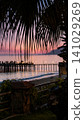 serene sunset over the beach with palm fronds and a wooden pier from a balcony overlooking the tranquil sea and sandy shore with warm pink and purple hues in the sky. 141029269