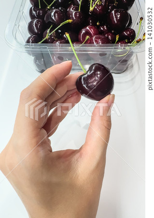 A hand holds a single cherry by its stem above a container. Fresh summer cherry for a healthy snack 141030532