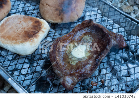 A juicy beef steak with melting butter on top and burger buns grilling over hot charcoal on a wire rack A juicy beef steak with melting butter on top and burger buns grilling over hot charcoal on a wire rack 141030742