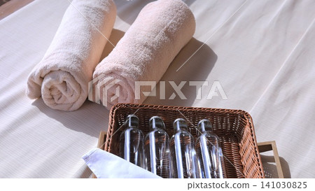 Rolled towels and clear bottles in 5 star hotel on wicker tray beside neutral linen in sunlit spa interior with soft shadows, calm minimal styling, warm texture, and hospitality comfort 141030825