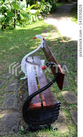 Ornamental park bench with swan shaped cast iron ends, wooden slats, painted details, and tropical garden setting beside stone pathway with lush greenery, with bench, swan, iron, wooden detail Ornamental park bench with swan shaped cast iron ends, wooden slats, painted details, and tropical garden setting beside stone pathway with lush greenery, with bench, swan, iron, wooden detail 141030826