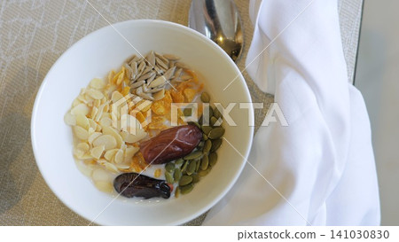 Top view breakfast bowl with milk soaked corn flakes, sliced almonds, sunflower seeds, pumpkin seeds and dates on linen placemat beside folded white napkin, neutral tabletop styling Top view breakfast bowl with milk soaked corn flakes, sliced almonds, sunflower seeds, pumpkin seeds and dates on linen placemat beside folded white napkin, neutral tabletop styling 141030830