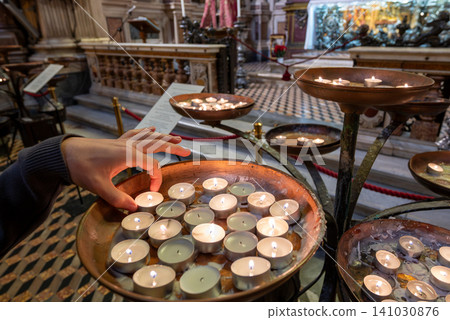 Naples, italy. December 27, 2024. Person's hand placing a lit votive tea light candle into a copper basin, praying for faith and devotion inside the cathedral of the assumption of mary Naples, italy. December 27, 2024. Person's hand placing a lit votive tea light candle into a copper basin, praying for faith and devotion inside the cathedral of the assumption of mary 141030876
