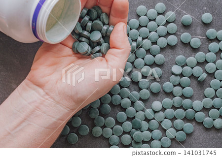 Close up top view of a person pouring round green spirulina or chlorella supplement tablets from a white plastic bottle container into an open hand Close up top view of a person pouring round green spirulina or chlorella supplement tablets from a white plastic bottle container into an open hand 141031745
