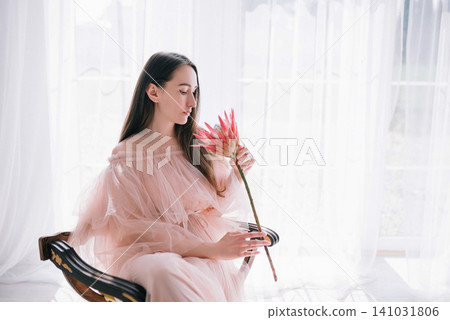 Elegant young woman and girl in pink tulle gown sitting in a classic chair by the window Elegant young woman and girl in pink tulle gown sitting in a classic chair by the window 141031806