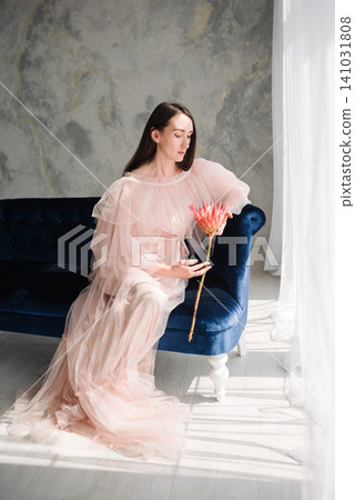 Elegant young woman and girl in pink tulle dress sitting on a blue sofa with a protea flower Elegant young woman and girl in pink tulle dress sitting on a blue sofa with a protea flower 141031808