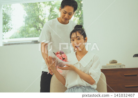 A scene depicting a couple where the girlfriend is surprised when her boyfriend presents her with a bouquet of pink roses indoors. A scene depicting a couple where the girlfriend is surprised when her boyfriend presents her with a bouquet of pink roses indoors. 141032407