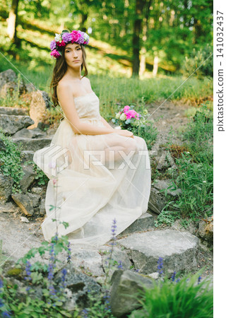 Young woman in flower wreath and beige dress sitting on stone steps in summer garden 141032437