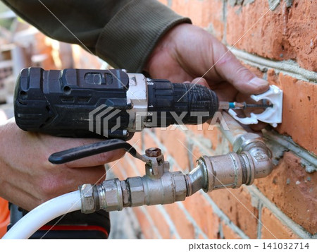 A technician uses a power screwdriver to fasten a white plastic clip to a brick wall with a self-tapping screw. In the foreground, beneath the tool, runs an assembled metal plumbing assembly featuring A technician uses a power screwdriver to fasten a white plastic clip to a brick wall with a self-tapping screw. In the foreground, beneath the tool, runs an assembled metal plumbing assembly featuring 141032714