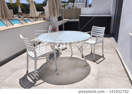 Modern outdoor patio with white table and chairs beside a swimming pool, featuring a sunny terrace and resort atmosphere, ideal for relaxing, dining, and enjoying a summer vacation Modern outdoor patio with white table and chairs beside a swimming pool, featuring a sunny terrace and resort atmosphere, ideal for relaxing, dining, and enjoying a summer vacation 141032782