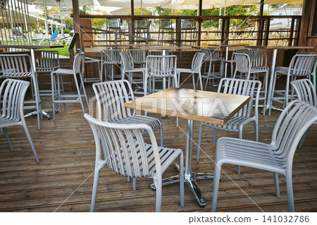 Modern outdoor restaurant terrace with wooden deck, stylish tables and chairs, creating a welcoming dining space for guests in a resort or cafe eon a sunny day. Modern outdoor restaurant terrace with wooden deck, stylish tables and chairs, creating a welcoming dining space for guests in a resort or cafe eon a sunny day. 141032786