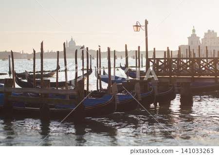 Traditional Venetian gondolas moored at a wooden pier in the lagoon during a hazy sunrise in Venice 141033263