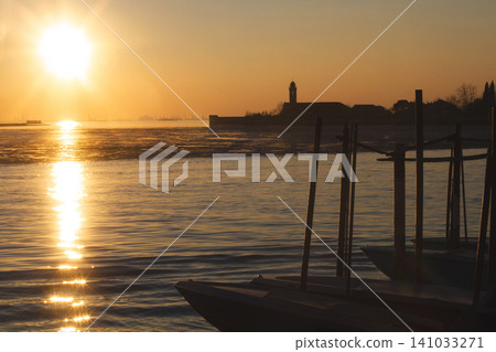 Golden sunset over the Venetian Lagoon with silhouettes of moored boats and a distant church Golden sunset over the Venetian Lagoon with silhouettes of moored boats and a distant church 141033271