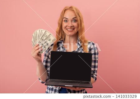 Smiling woman with red hair showing her earnings, holding a fan of dollar cash in one hand and a blank screen laptop in the other, celebrating online financial success and digital income 141033329