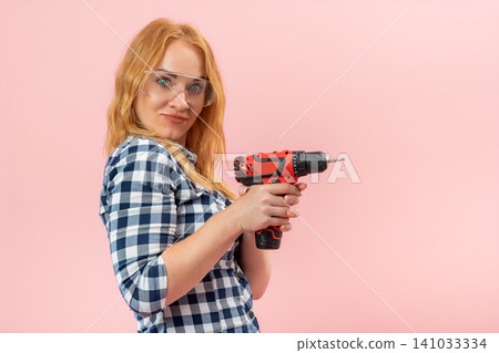 Young woman in plaid shirt and safety glasses holding a power drill, confidently looking at the camera, embodying female empowerment and readiness for home improvement projects Young woman in plaid shirt and safety glasses holding a power drill, confidently looking at the camera, embodying female empowerment and readiness for home improvement projects 141033334