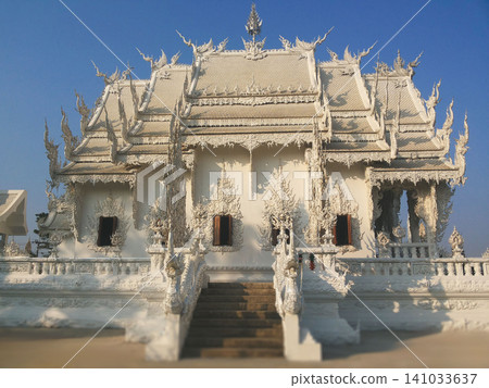 Front view of the famous White Temple featuring traditional Thai tiered roof and detailed mirror glass mosaics under a clear blue sky. Front view of the famous White Temple featuring traditional Thai tiered roof and detailed mirror glass mosaics under a clear blue sky. 141033637